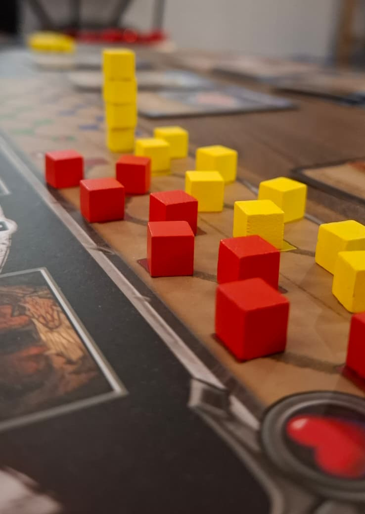 Close-up of the corner of a board game board, on a dark table.  There are small coloured wooden cubes in two zigzag lines, one red and one yellow.  The yellow line is longer and turns into a vertical stack of cubes at the end.  A board game fan might recognise it as the game Clank! at or very near the end, after the yellow player has died.