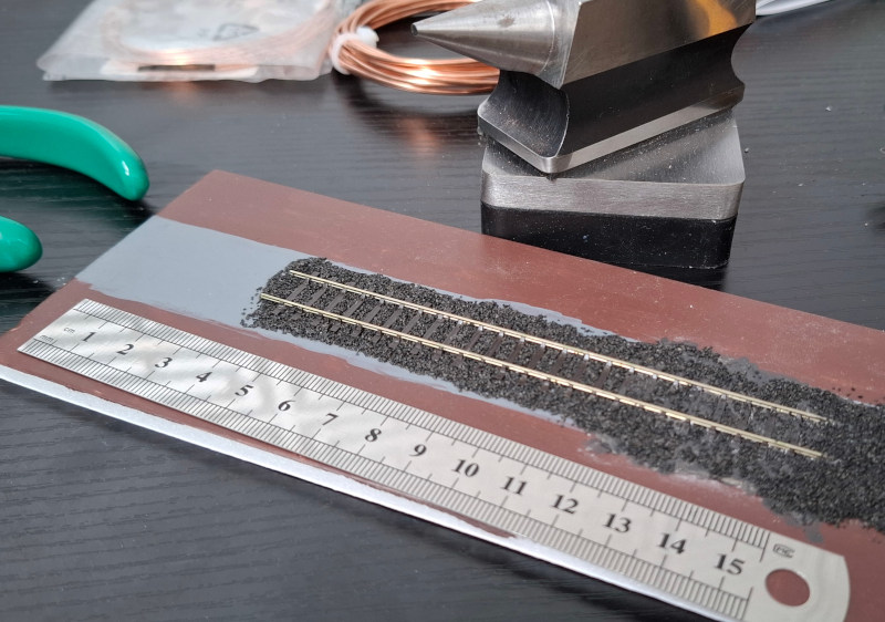 A piece of model train track, stuck down to a small piece of painted foamboard, on the same workbench as the last picture and next to the same ruler. You can see that the track gauge is roughly the same, but the rails are much smaller and thinner, and the sleepers are spaced more widely apart.
