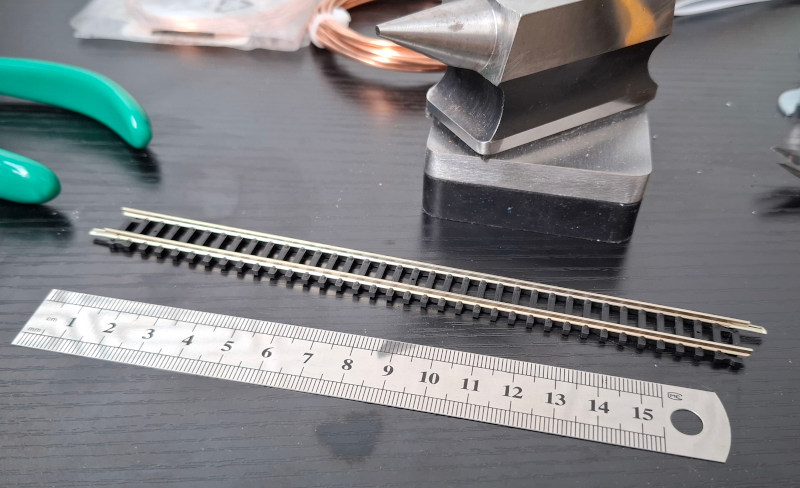 A piece of model train track on a black workbench, next to a 15cm steel ruler. You can see, by comparing the ruler and the track, that the track gauge is around nine millimetres. The track is quite chunky, with thick rails and sleepers.
